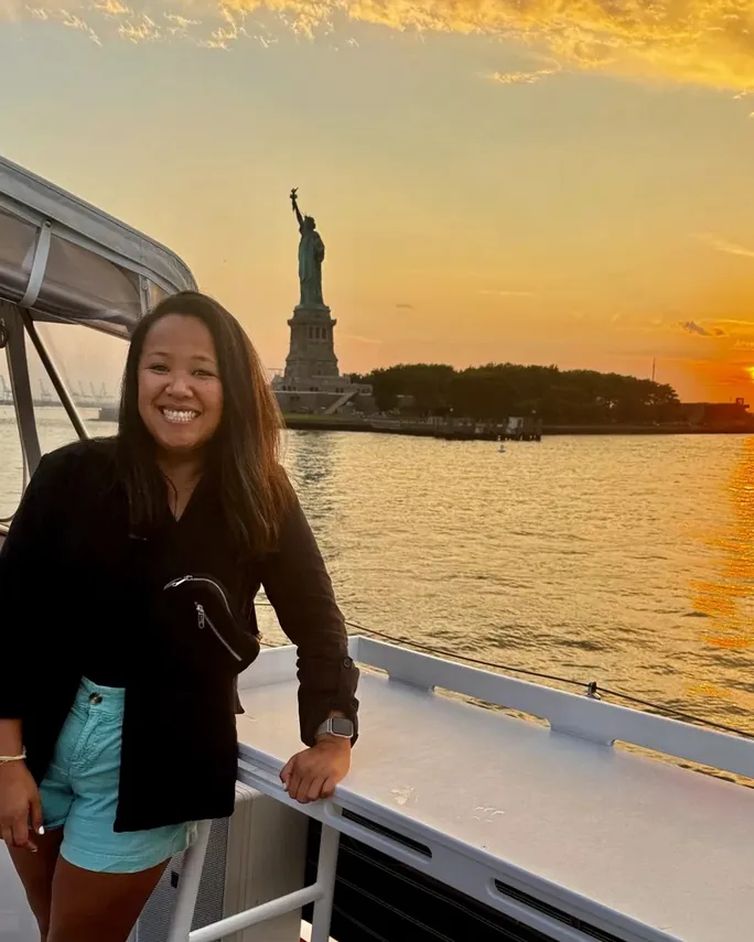 Corina Violette smiling on a boat at sunset, the Statue of Liberty behind her